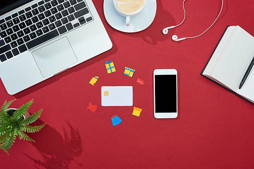 top view of credit card with icons on red background with smartphone, laptop, earphones, coffee, notebook and plant