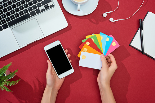 cropped view of woman holding multicolored empty credit cards and smartphone on red background with laptop, earphones and coffee
