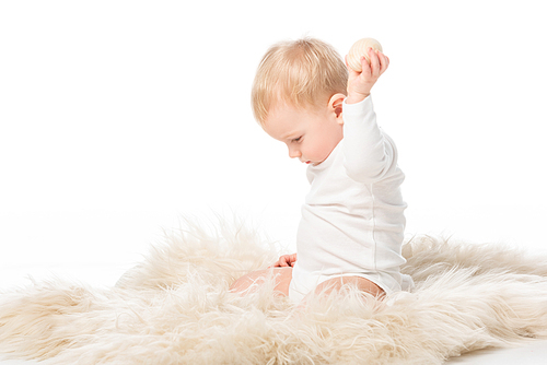 side view of cute kid looking down and holding . egg in raised hand, sitting on fur on white background
