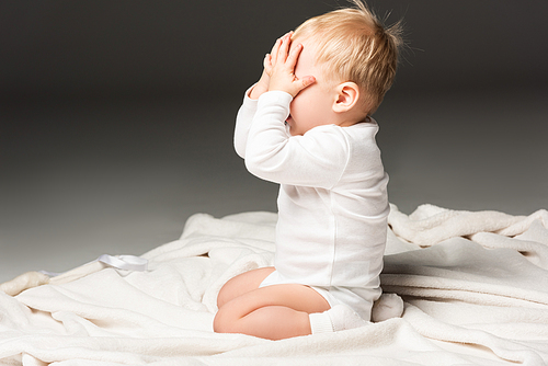 Side view of cute child covering face and kneeling on blanket isolated on black