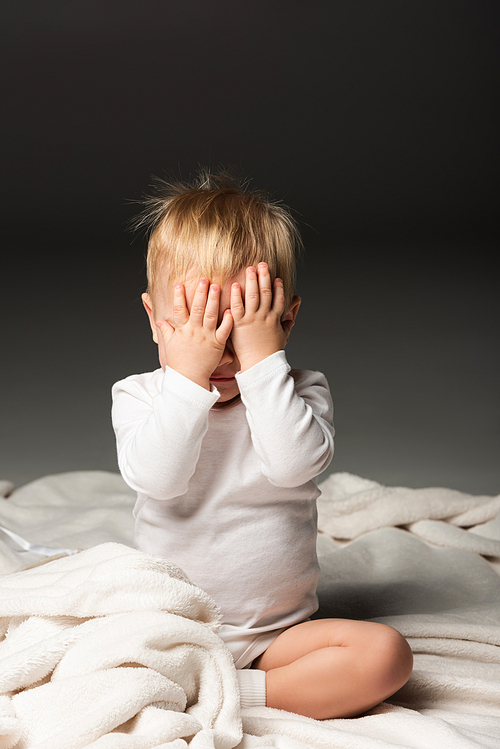 Cute child covering face with hands and sitting on blanket on black background