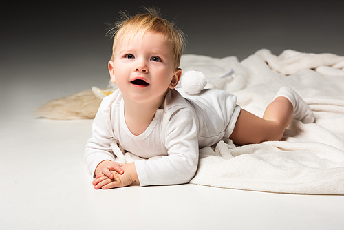Child with pompon on underpants, lying on blanket, looking up with open mouth on grey background