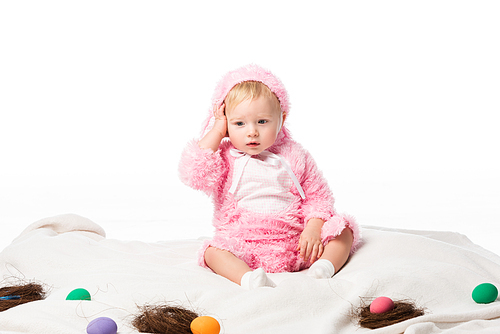 Upset child wearing rabbit costume, touching head on blanket with colorful decoration isolated on white