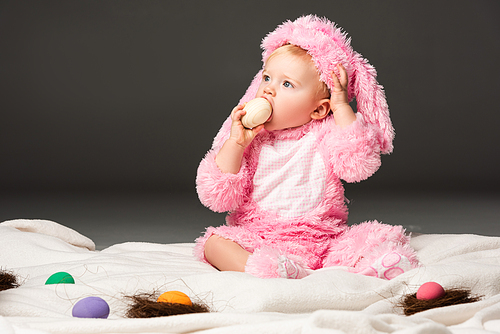 child wearing rabbit costume, putting . egg in mouth and sitting on blanket isolated on black