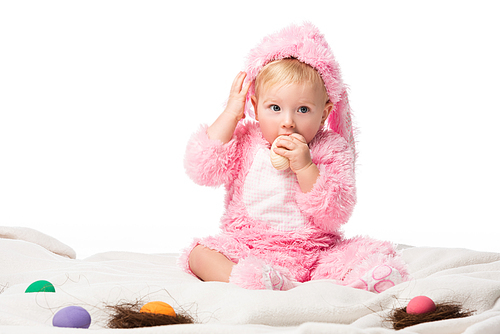 Child wearing rabbit costume, putting easter egg in mouth, touching head on blanket isolated on white