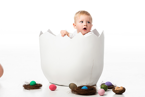 Cute child with open mouth inside eggshell with nests and easter eggs on white background