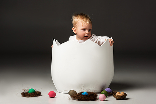 Child with open mouth holdind eggshell while sitting inside on black background