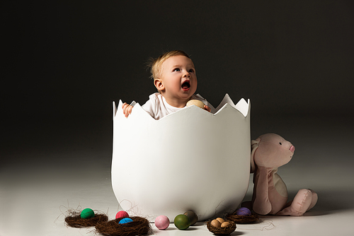 child with open mouth looking up, holding . egg in eggshell on black background