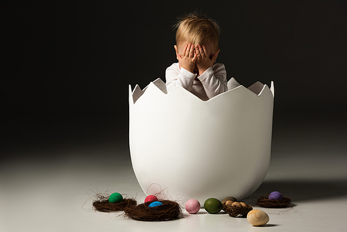 child covering face inside eggshell next to . eggs and nests on black background