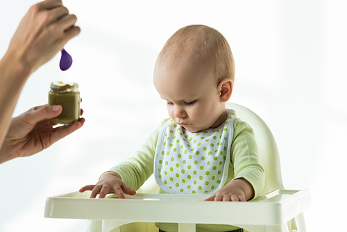 Selective focus of mother holding jar of baby nutrition near baby son on feeding chair on white background