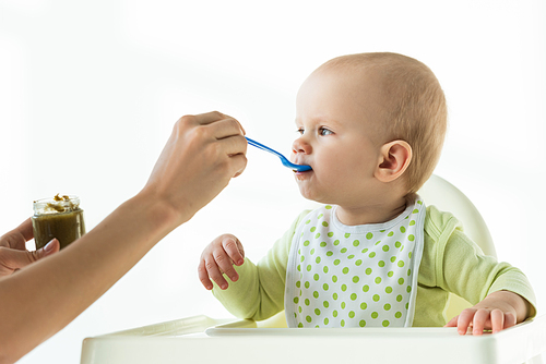 Mother with jar of baby nutrition and spoon feeding infant on feeding chair on white background