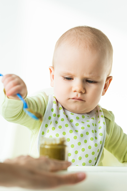 Selective focus of baby holding spoon near jar with vegetable puree and mother on white background