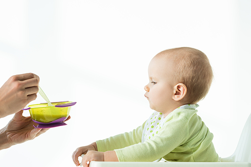 Side view of mother holding bowl of baby nutrition and spoon near infant on feeding chair on white background
