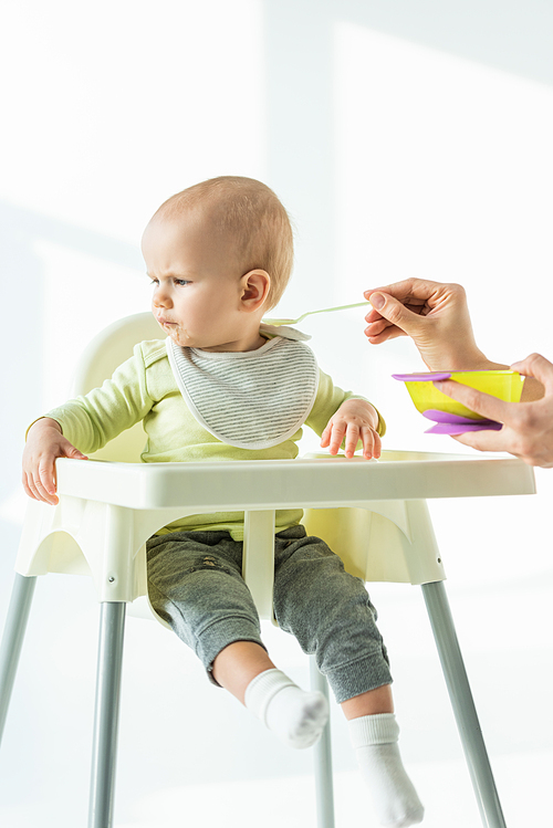 Baby on feeding chair looking away near mother with bowl of baby nutrition and spoon on white background