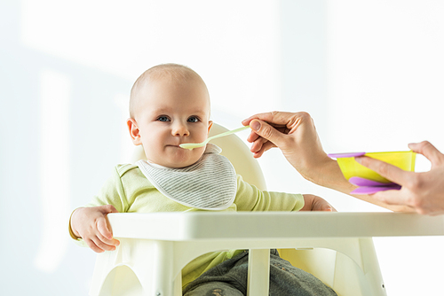 Selective focus of mother with puree feeding baby on feeding chair on white background