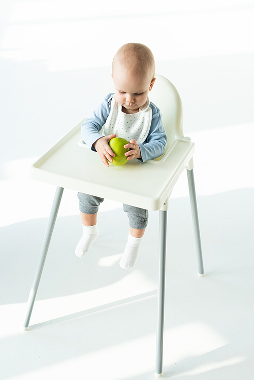 Cute baby holding ripe apple while sitting on feeding chair on white background