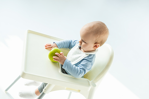 High angle view of baby boy holding green apple while sitting on feeding chair on white background