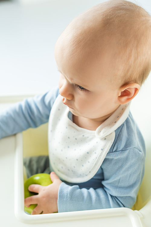 Side view of baby boy holding green apple while sitting on feeding chair on white background