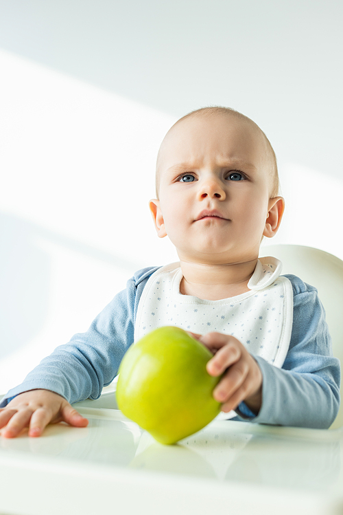 Selective focus of pensive baby boy holding green apple while sitting on feeding chair on white background