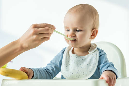 Mother feeding baby son while holding banana on table of feeding chair on white background