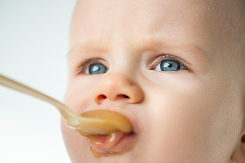 Close up view of adorable baby boy looking away during feeding with fruit puree isolated on grey