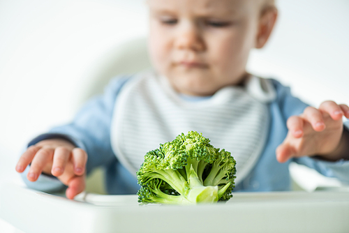 Selective focus of piece of broccoli on table near baby on feeding chair isolated on grey