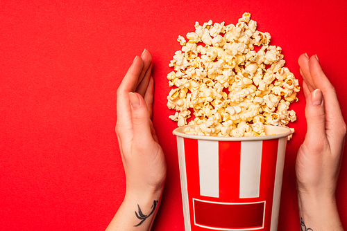 Top view of female hands near bucket with tasty popcorn on red background