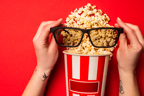 Top view of girl holding sunglasses near cardboard bucket with popcorn on red background