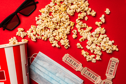 Top view of popcorn, medical mask with cinema tickets and sunglasses on red background