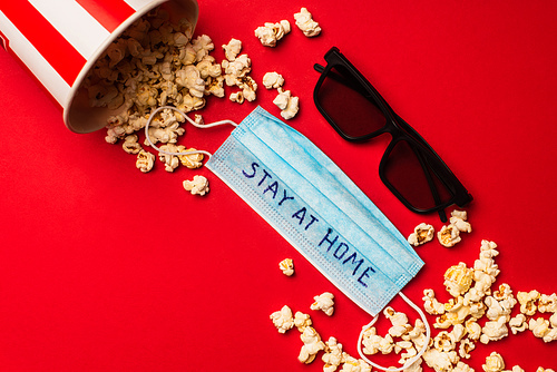 Top view of medical mask with stay at home lettering near sunglasses and popcorn on red background