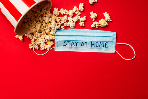 Top view of bucket with popcorn and medical mask with stay at home lettering on red background