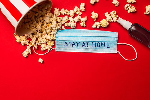 Top view of medical mask with stay at home lettering near hand sanitizer and popcorn on red background