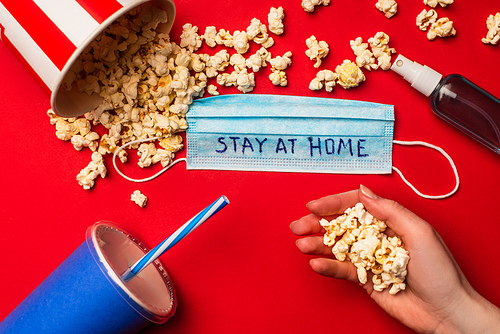 Top view of woman holding popcorn near medical mask with stay at home lettering, hand sanitizer and paper cup on red background