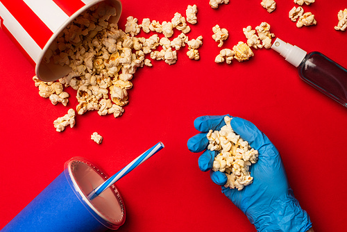 Top view of person in latex glove holding popcorn near hand sanitizer and paper cup on red background