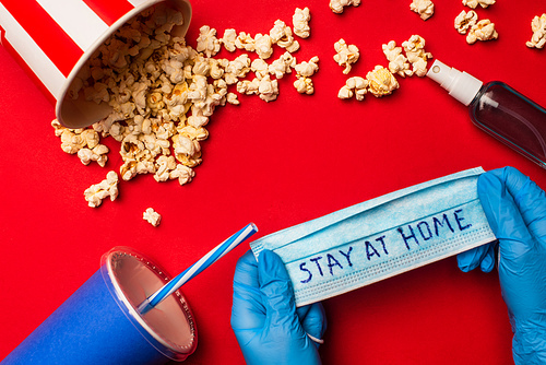 Top view of person holding medical mask with stay at home lettering near hand sanitizer and popcorn on red surface
