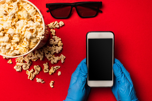 Top view of person holding smartphone with blank screen near sunglasses and popcorn on red background
