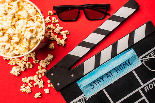 Top view of clapperboard with medical mask and stay at home lettering near popcorn and sunglasses on red surface