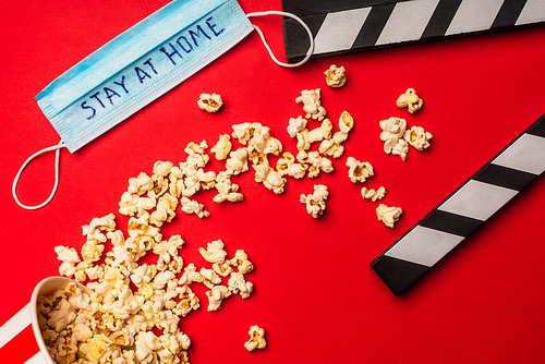 Top view of popcorn near clapperboard and medical mask with stay at home lettering on red background