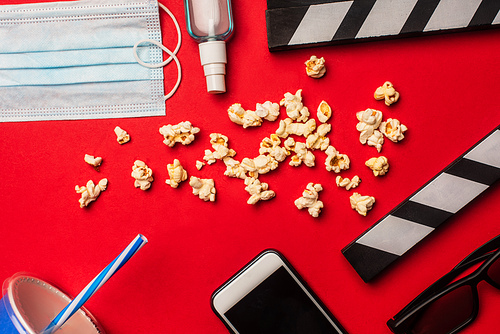 Top view of medical mask, smartphone with hand sanitizer and popcorn on red background