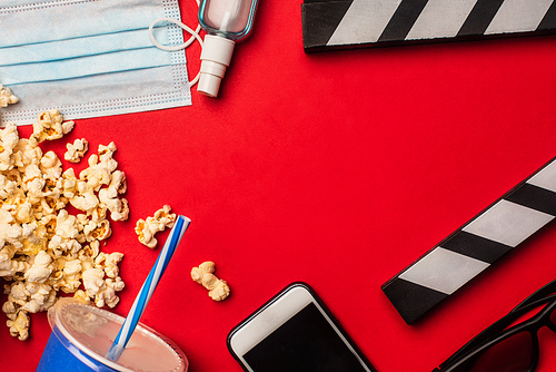 Top view of popcorn, smartphone with medical mask and hand sanitizer on red surface