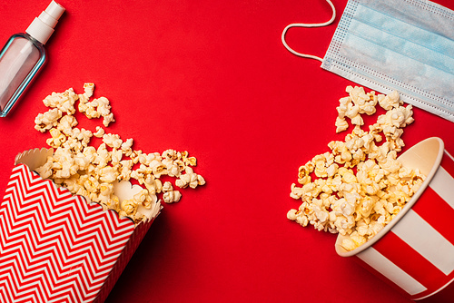 Top view of hand sanitizer, medical mask and buckets with popcorn on red surface