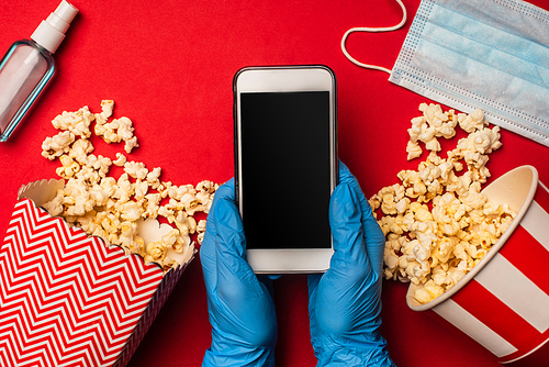 Top view of person holding smartphone with blank screen near medical mask and popcorn on red surface