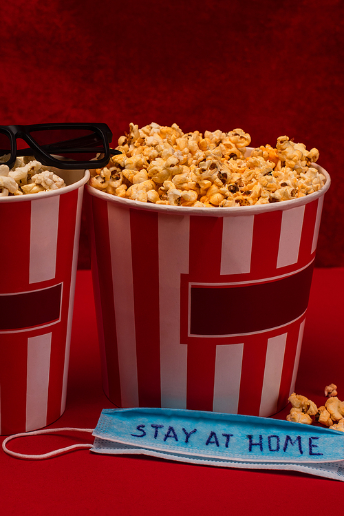 Medical mask with stay at home lettering near buckets with popcorn and sunglasses on red surface and velour at background