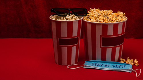 Buckets with popcorn near sunglasses and medical mask with stay at home lettering on red surface and velour at background