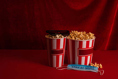 Buckets with popcorn near medical mask with stay at home lettering and sunglasses on red surface with velour at background