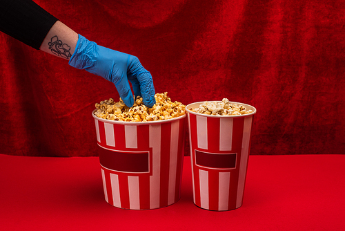 Cropped view of woman in latex glove taking popcorn from bucket on red surface with velour at background