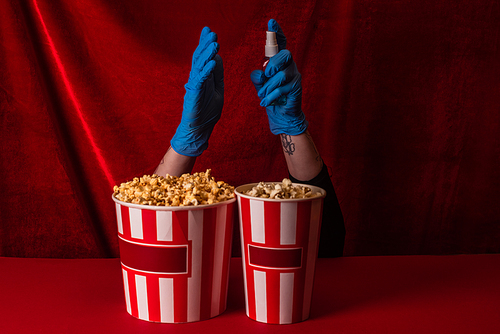 Cropped view of woman in latex gloves holding hand sanitizer near buckets with popcorn on red surface with velour at background
