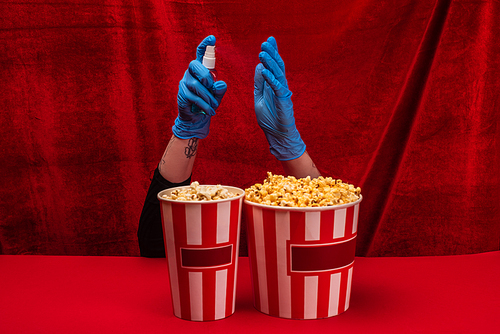 Cropped view of woman in latex gloves holding bottle of hand sanitizer near popcorn on red surface with velour at background