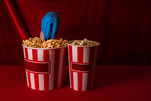 Cropped view of woman in latex glove taking popcorn from bucket on red surface and velour at background