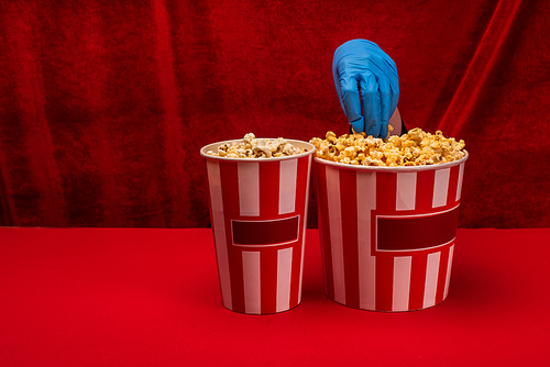 Cropped view of woman in latex glove taking popcorn from bucket on red surface with velour at background